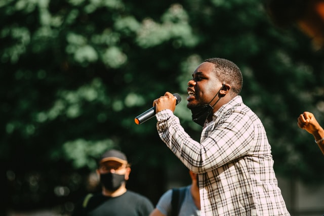 A man in a plaid shirt speaking wholeheartedly in public depicts how to be a better public speaker. 